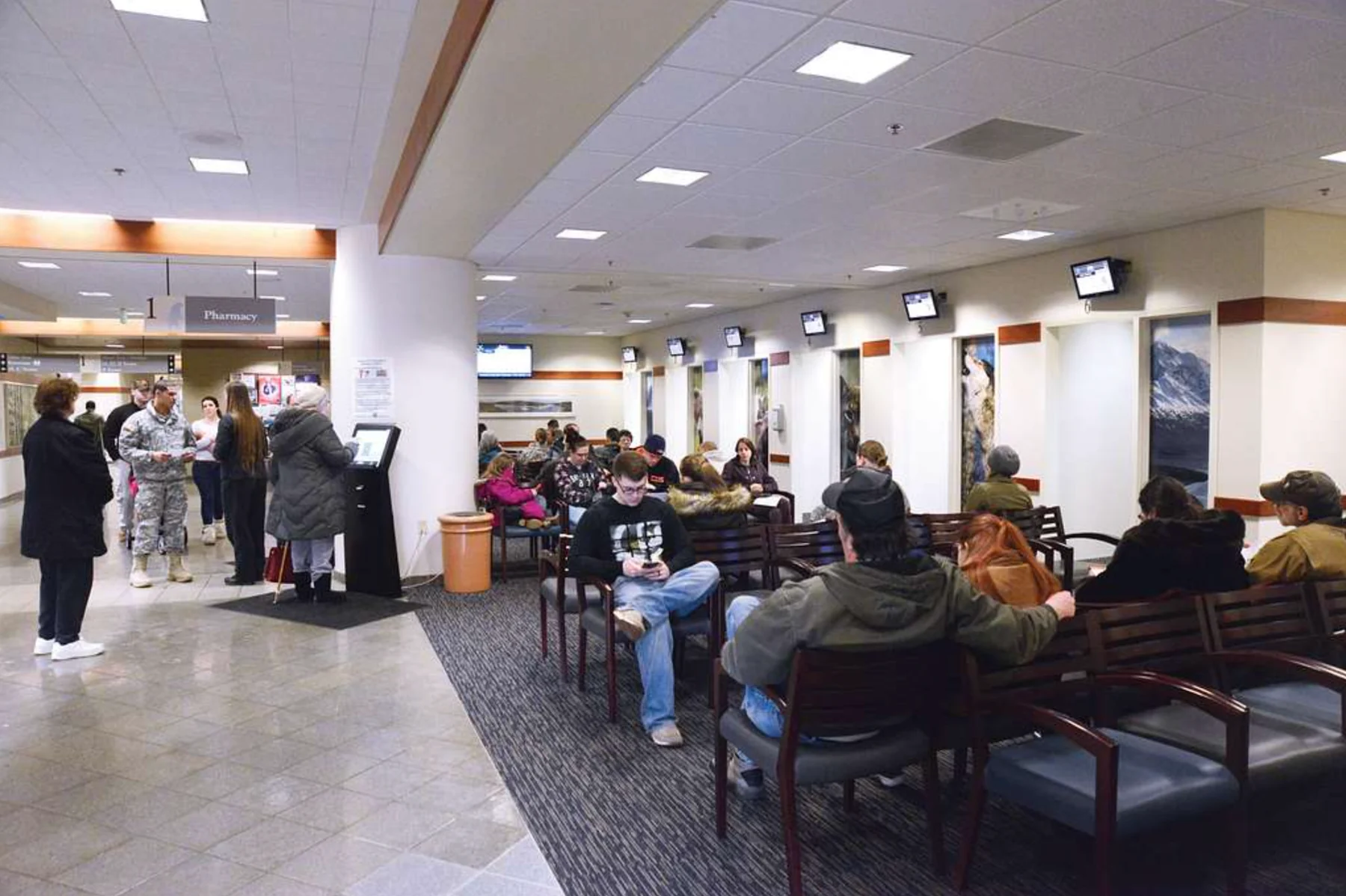 Hospital waiting room showing patients, staff, kiosks, seating, signage, and information displays.