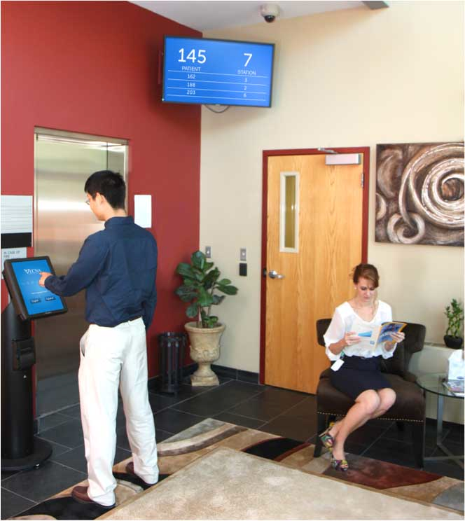 A person using a VetLink kiosk while a patient waits beneath a queue display